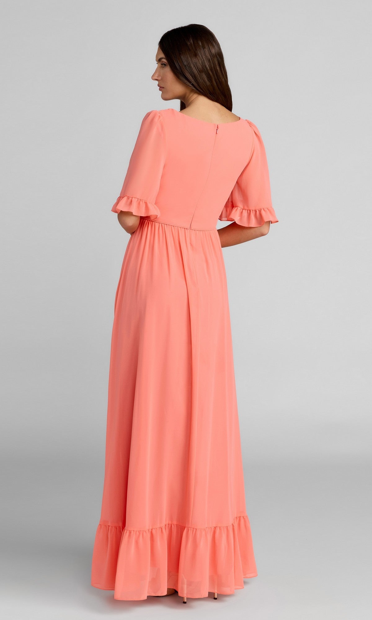 A woman stands facing away, wearing the Mac Duggal Long Formal Dress 55867 in coral-pink with short ruffled sleeves and a ruffled hem, set against a plain gray background.